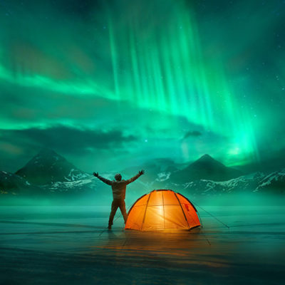 A man camping in wild northern mountains with an illuminated tent viewing a spectacular green northern lights aurora display. Photo composition.